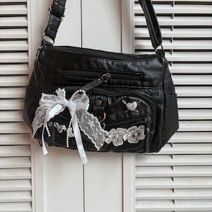 Puede incluir: Bolso bandolera negro con múltiples compartimentos con cremallera. El bolso está decorado con un lazo de encaje blanco, detalles florales y un alfiler en forma de corazón. Tiene una correa larga ajustable.