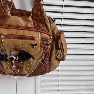 May include: A tan and brown handbag with multiple pockets and decorative charms. The bag features a bat wing, cross, and butterfly embellishments, along with safety pins and chains. The bag has a zipper closure and gold-colored hardware.