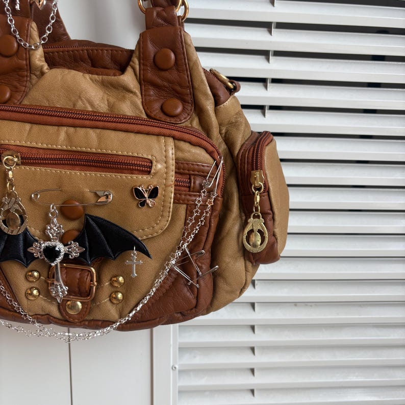May include: A tan and brown handbag with multiple pockets and decorative charms. The bag features a bat wing, cross, and butterfly embellishments, along with safety pins and chains. The bag has a zipper closure and gold-colored hardware.