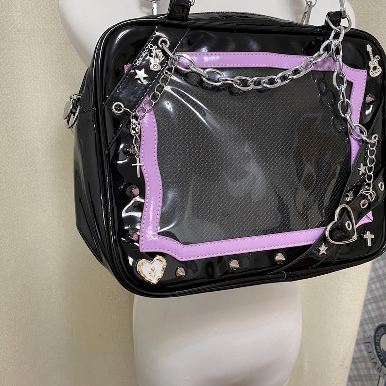 Puede incluir: Bolso de mano de charol negro con una ventana de pl&aacute;stico transparente y ribete morado. El bolso tiene una correa de cadena plateada y detalles met&aacute;licos.