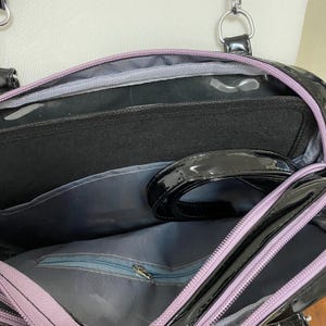 Puede incluir: El interior de un bolso negro y morado con un bolsillo con cremallera y una correa negra. El bolso tiene un acabado brillante de charol.