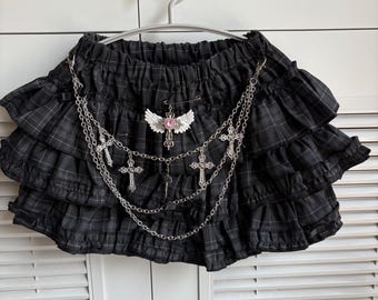 Puede incluir: Falda a cuadros negros con volantes en capas, colgada de una percha. La falda presenta una cadena plateada con dijes de cruz y un alfiler decorativo con alas y una gema rosa.