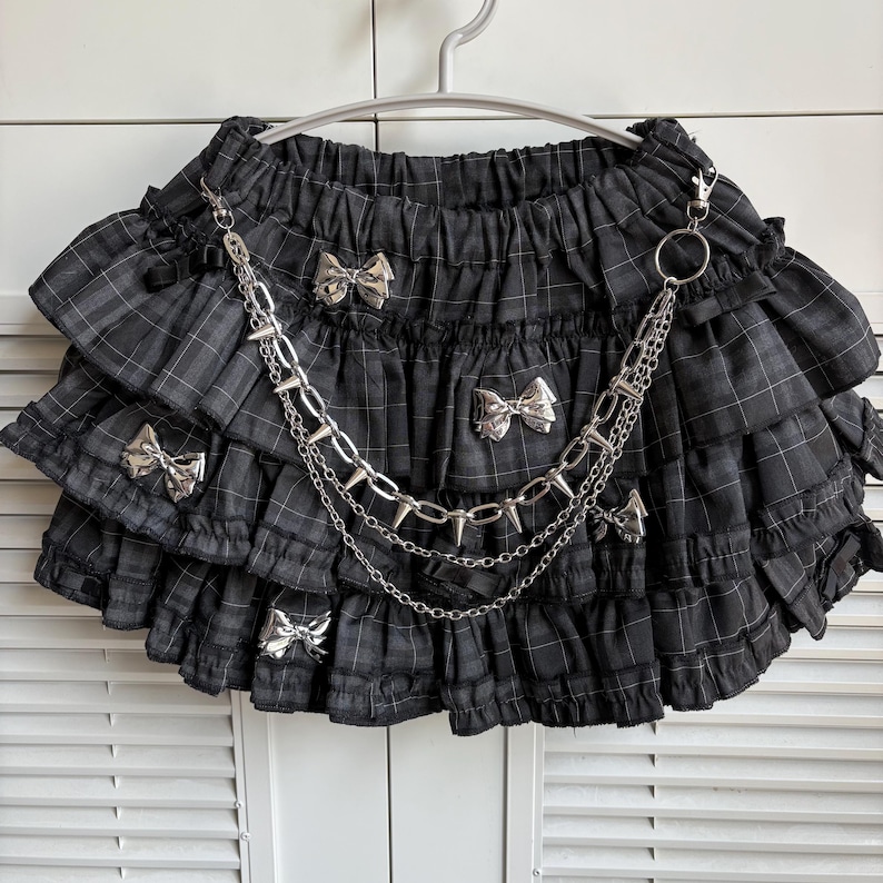 May include: Black plaid skirt with multiple ruffled layers, adorned with silver bow accents and a silver chain with spikes. The skirt is hanging on a white hanger.
