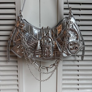 Puede incluir: Bolso plateado con textura de cocodrilo, adornado con púas plateadas, cadenas y un cierre de hebilla. El bolso tiene una correa para el hombro y un pequeño bolsillo con un detalle de perla. El diseño es atrevido y elegante.