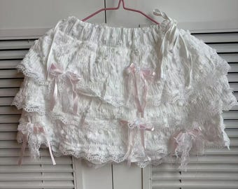 May include: White ruffled skirt with multiple layers and delicate lace trim. The skirt features several pink satin bows and is hanging on a pink hanger. The fabric has a textured, almost quilted appearance.