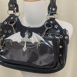 May include: A black patent leather handbag with a clear plastic front panel. The bag has silver studs, grommets, and chains. A silver cross with a white bat wing charm hangs from the bag.