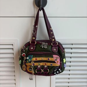 May include: A dark olive green handbag with purple trim, featuring a front zippered pocket, metallic studs, colorful rhinestones, and silver safety pins attached to the handles and body. The bag has two handles and a multi-textured exterior design.