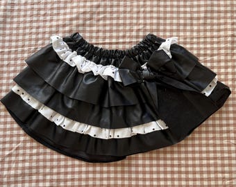 以下が含まれることがあります： 黒のフェイクレザー製スカート。白い縁取りと小さな黒い水玉模様があしらわれた段状のフリルが特徴です。ウエストはゴム仕様で、右側には大きな黒いリボンが取り付けられており、重なり合ったデザインになっています。