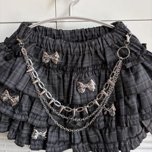 May include: A black and gray plaid skirt with multiple ruffled layers. The skirt features silver bow accents and a silver chain with spikes. The skirt is hanging on a white hanger.