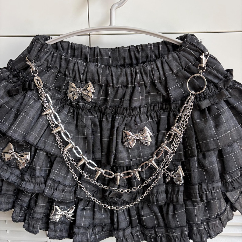 May include: A black and gray plaid skirt with multiple ruffled layers. The skirt features silver bow accents and a silver chain with spikes. The skirt is hanging on a white hanger.