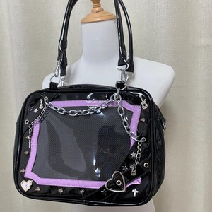 Puede incluir: Bolso de mano de charol negro con un panel de pl&aacute;stico transparente y detalles de cadena plateada. El bolso tiene un ribete morado y presenta un coraz&oacute;n, estrellas y cruces.
