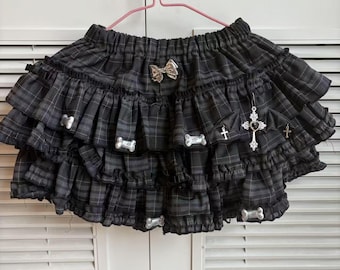 May include: A layered, ruffled skirt in a dark gray plaid pattern. The skirt is adorned with silver-colored bone and cross charms, and a bow. The skirt is hanging on a pink hanger.