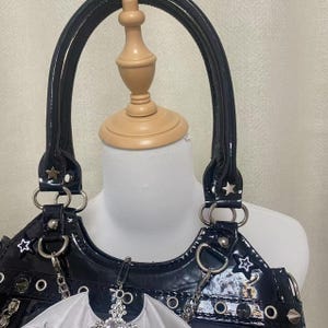 May include: A black patent leather handbag with silver star accents and a silver chain strap. The bag has a white bat wing charm attached to the front.