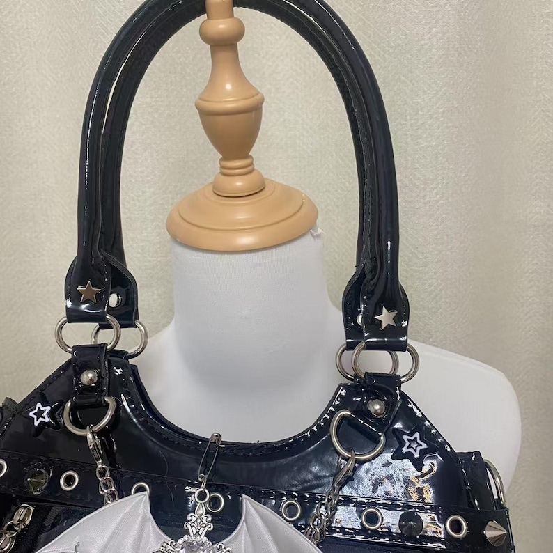 May include: A black patent leather handbag with silver star accents and a silver chain strap. The bag has a white bat wing charm attached to the front.