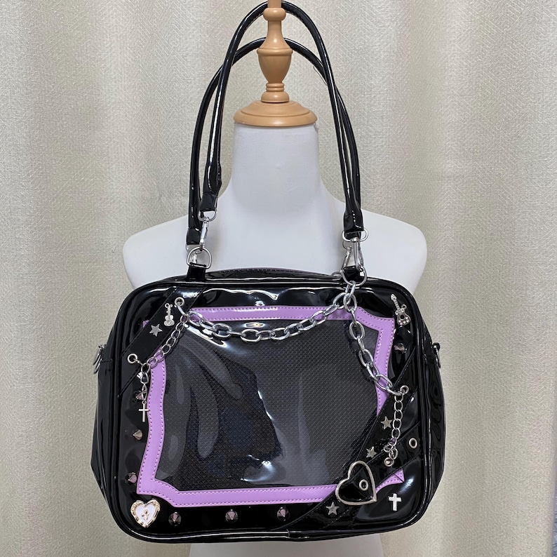 Puede incluir: Bolso de mano de charol negro con una ventana de pl&aacute;stico transparente y detalles de cadena plateada. El bolso tiene un ribete morado y presenta un dise&ntilde;o de coraz&oacute;n y cruz.