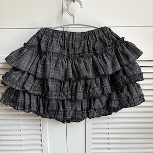 May include: A gray plaid ruffled skirt hanging on a silver hanger. The skirt has multiple layers of ruffles and a gray and white plaid pattern. The skirt is made of a soft fabric.