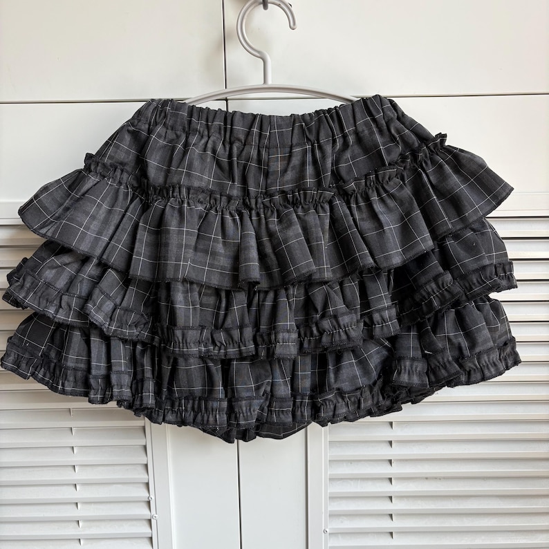May include: A gray plaid ruffled skirt hanging on a silver hanger. The skirt has multiple layers of ruffles and a gray and white plaid pattern. The skirt is made of a soft fabric.