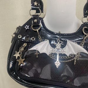 May include: A black patent leather handbag with a clear plastic front panel. The bag has silver star and cross accents, and a silver chain strap. A white bat wing charm hangs from the bag.