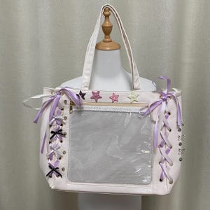 May include: A cream-coloured tote bag with a clear front pocket and a zip closure. The bag is decorated with lilac ribbons, black bows, and star-shaped embellishments. It has two handles and a shoulder strap.