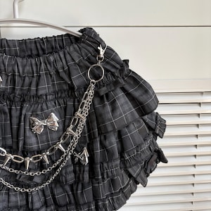 May include: Black and gray plaid skirt with multiple ruffled layers. The skirt features silver bow accents and a silver chain with spikes. The skirt is hanging on a white hanger.