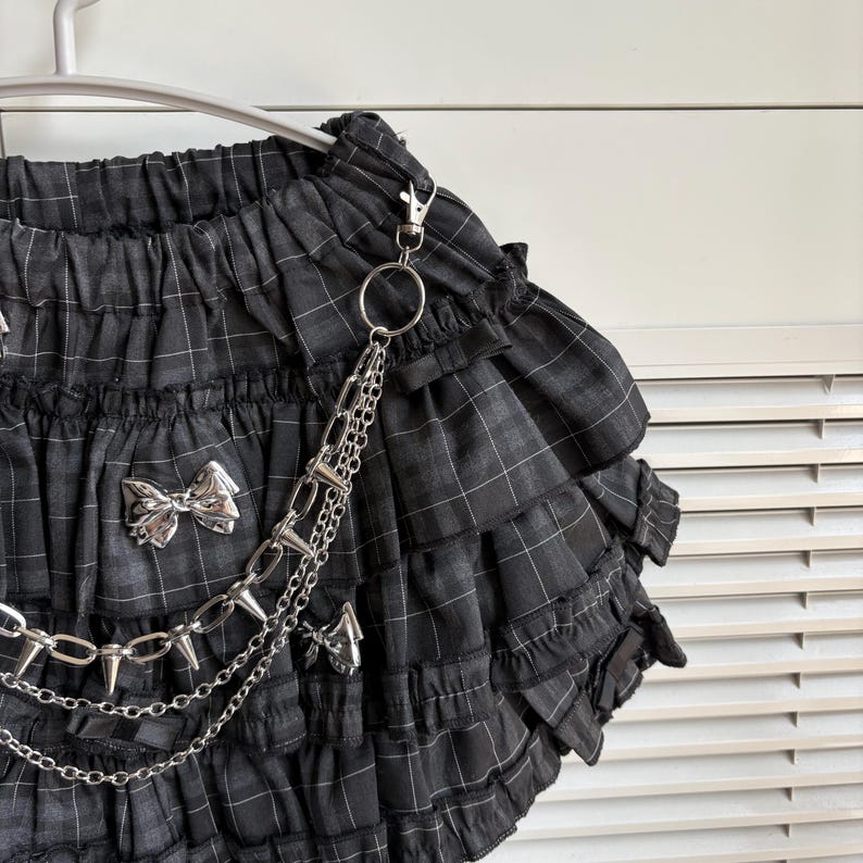 May include: Black and gray plaid skirt with multiple ruffled layers. The skirt features silver bow accents and a silver chain with spikes. The skirt is hanging on a white hanger.