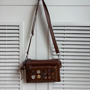 May include: A brown crossbody bag with a long strap, hanging against a white shuttered door. The bag features multiple zippers and a front pocket adorned with various buttons and charms. The bag is made of a leather-like material.