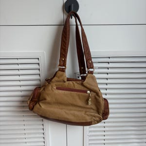 May include: A tan handbag with brown accents and straps hangs from a black hook. The bag features a zippered main compartment, side pockets, and a gold-colored zipper pull. The bag's design includes a combination of textures and colors.