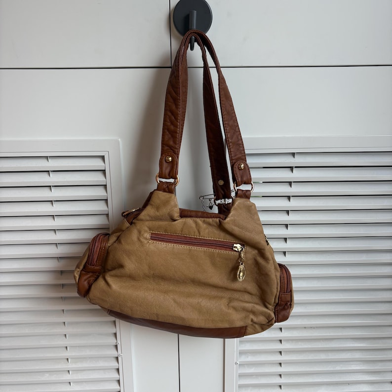 May include: A tan handbag with brown accents and straps hangs from a black hook. The bag features a zippered main compartment, side pockets, and a gold-colored zipper pull. The bag's design includes a combination of textures and colors.