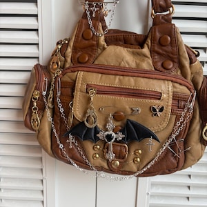May include: A brown and tan handbag with multiple pockets and decorative metal accents. The bag features a black bat wing applique and silver chain details with charms. The bag has a zipper closure and a buckle.