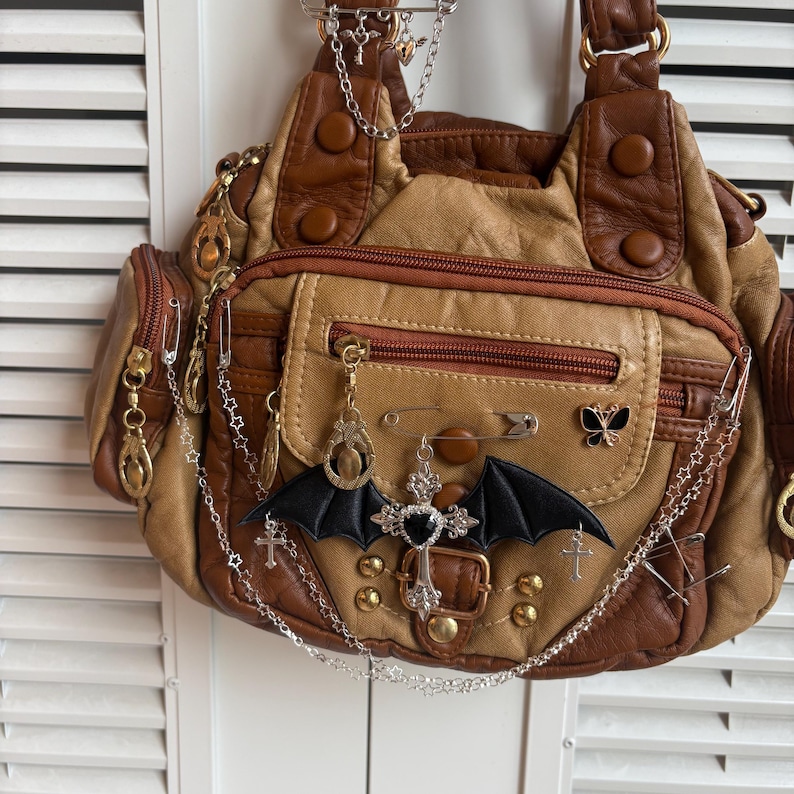May include: A brown and tan handbag with multiple pockets and decorative metal accents. The bag features a black bat wing applique and silver chain details with charms. The bag has a zipper closure and a buckle.