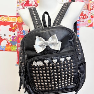 May include: A black faux leather backpack featuring a large silver metallic bow on the front pocket. The pocket is decorated with rows of silver pyramid studs, four white heart-shaped accents, and a silver bat-shaped charm with clear crystals.
