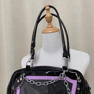 Puede incluir: Bolso de mano de charol negro con un panel de malla transparente y detalles de cadena plateada. El bolso tiene un ribete lavanda y un asa superior.