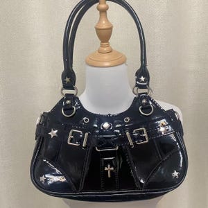 May include: Black patent leather handbag with silver hardware, including stars and a cross. The bag has a top handle and a buckle detail.