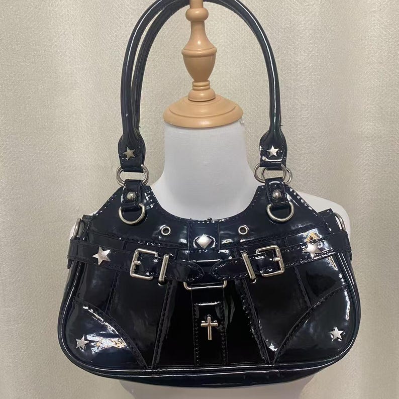 May include: Black patent leather handbag with silver hardware, including stars and a cross. The bag has a top handle and a buckle detail.