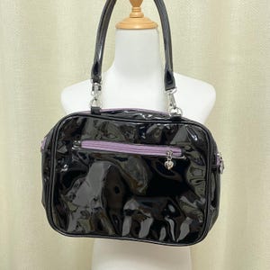 Puede incluir: Un bolso de mano de charol negro con una cremallera morada y un dije de coraz&oacute;n plateado.
