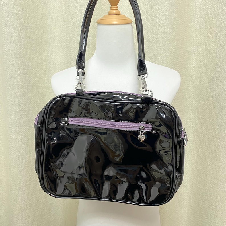 Puede incluir: Un bolso de mano de charol negro con una cremallera morada y un dije de coraz&oacute;n plateado.