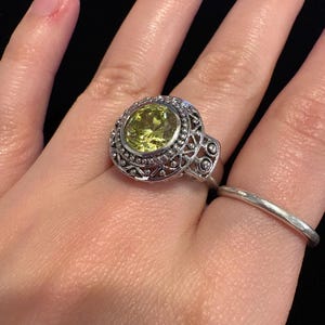 May include: A silver ring with an oval, light green gemstone set in an ornate, filigree design. The ring is on a hand, with a simple silver band on an adjacent finger. The gemstone is the focal point, surrounded by intricate metalwork.