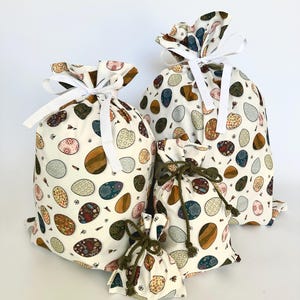 May include: Four fabric gift bags with a white background and a pattern of colourful eggs. The bags are different sizes and have white and brown drawstrings.