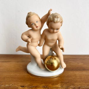 Puede incluir: Una figura de porcelana que representa a dos querubines jugando con una bola dorada. Las figuras desnudas tienen rasgos faciales detallados y están posadas sobre una base blanca con adornos dorados. La figura está sobre un fondo neutro.