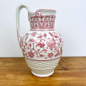 Könnte beinhalten: Eine weiße Keramikkrug mit einem roten Blumenmuster und einem geflochtenen Korbmuster am Boden. Der Krug hat einen gebogenen Griff und einen dekorativen Rand. Das Blumenmuster umfasst Rosen und Weinreben.