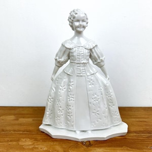 May include: A white porcelain figurine of a woman in a long dress with floral detailing. The figurine has a detailed bodice, puffed sleeves, and a lace collar. The figure stands on a hexagonal base.