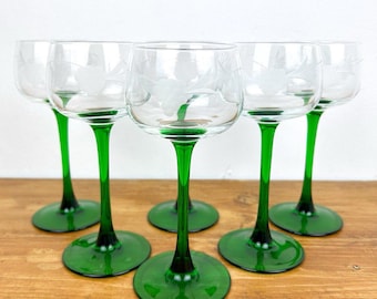 May include: Set of six clear wine glasses with etched floral designs. Each glass features a vibrant green stem and base. The glasses are arranged on a wooden surface, showcasing their elegant design and color contrast.