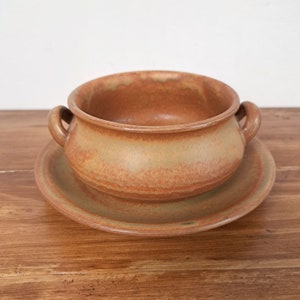 May include: A handmade, rustic ceramic soup bowl with two handles, resting on a matching saucer. The bowl and saucer are a warm, earthy brown with subtle variations in color, suggesting a natural, handcrafted aesthetic. The set is ideal for serving soups or stews.