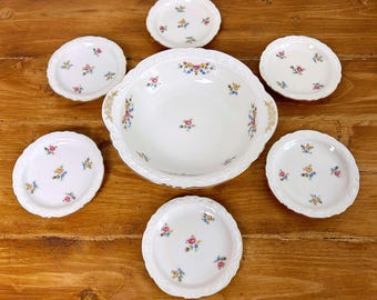 May include: A white porcelain serving set with a large bowl and six smaller plates. Each piece is decorated with a delicate floral pattern in pink, yellow, and blue. The set is arranged on a wooden surface.