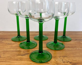 May include: Six clear wine glasses with emerald green stems and bases. The glasses are arranged on a wooden surface, showcasing their elegant design. The green stems provide a striking contrast to the clear glass bowls.