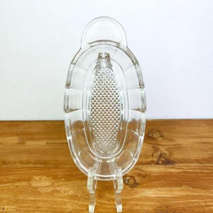 May include: Clear glass serving dish with an oval shape and a textured diamond pattern in the center. The dish has a handle and is displayed on a clear stand. The dish is likely vintage and suitable for serving appetizers or small portions.