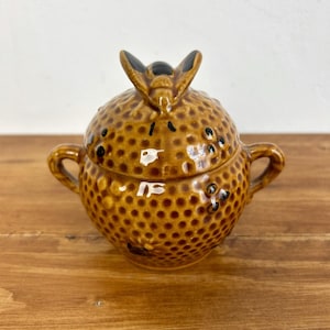 Può includere: Una zuccheriera in ceramica marrone con coperchio, caratterizzata da una superficie strutturata e due piccole maniglie. Il coperchio è adornato da un elemento decorativo. La ciotola è posta su una superficie di legno.