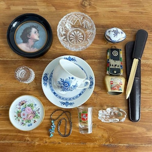 May include: A collection of vintage items including a small framed portrait, a crystal bowl, a teacup and saucer with blue floral designs, a small trinket box, a decorative bell, a letter opener, a shot glass, a small plate with floral design, a necklace, and a crystal ashtray.