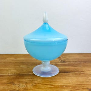 May include: A light blue glass candy dish with a lid, set on a white stem and base. The dish has a rounded bowl shape and a conical lid with a white tip. The base is circular, and the dish sits on a wooden surface.