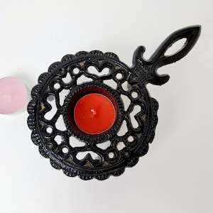 Könnte beinhalten: Schwarzer Kerzenhalter aus Gusseisen mit einem dekorativen, durchbrochenen Design. Der Halter hat eine zentrale Tasse mit einer brennenden roten Kerze. Eine rosa Kerze befindet sich daneben. Der Halter hat einen Griff.